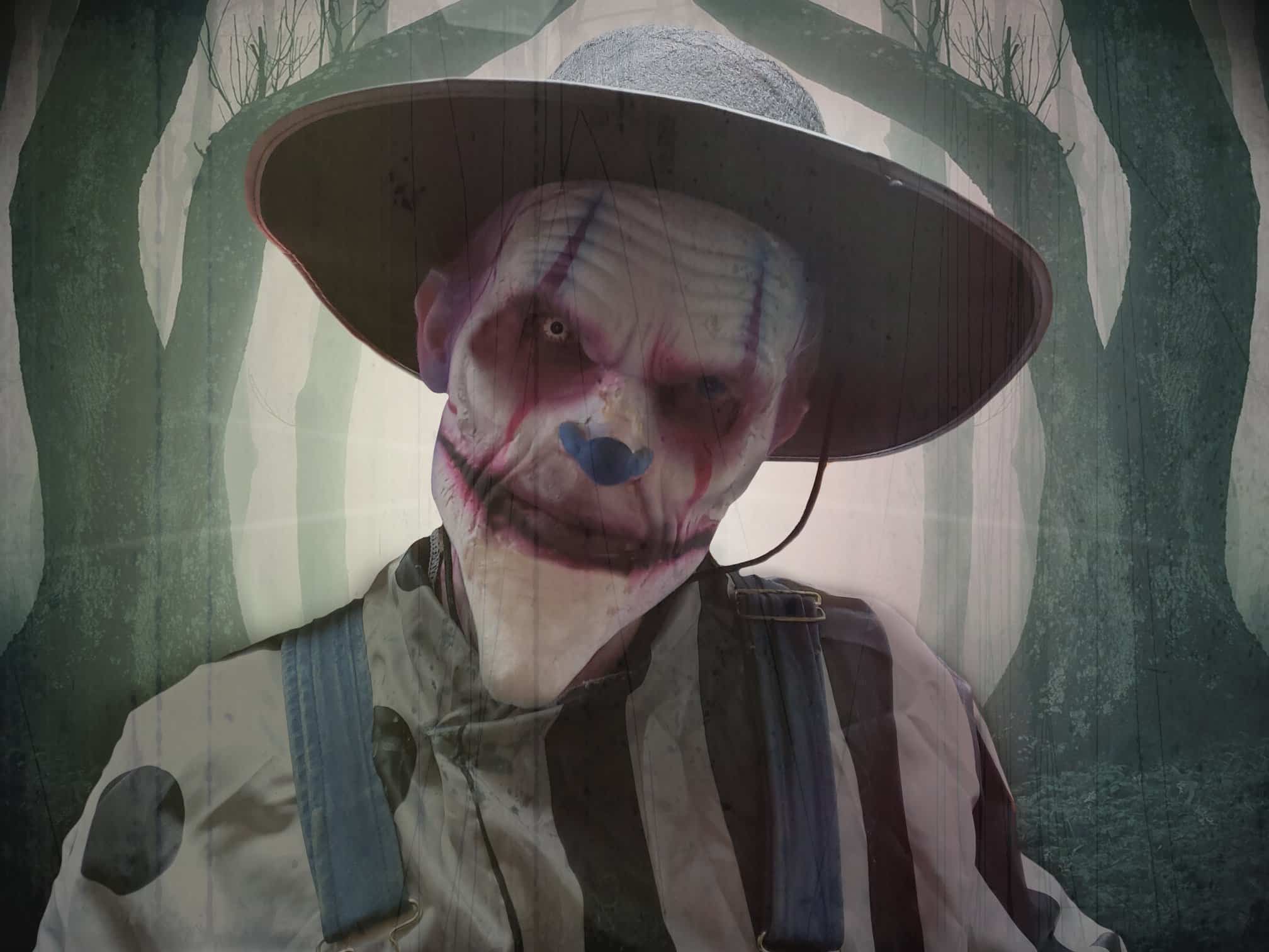 A Field of Screams character in a wide-brimmed hat and painted face standing in the dark woods.